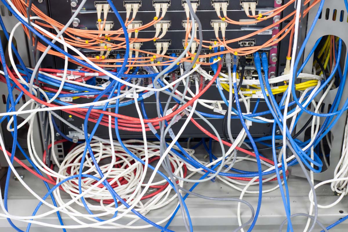 A close-up of a disorganized "spaghetti" server closet with tangled blue, red, and orange cables, illustrating neglected technical debt.