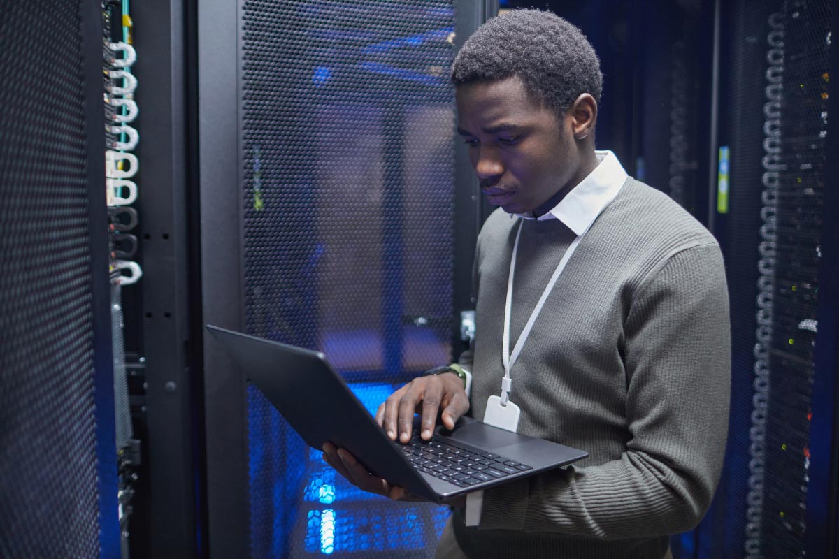 An IT specialist with a laptop performing a site-level discovery audit inside a server room to verify infrastructure readiness.